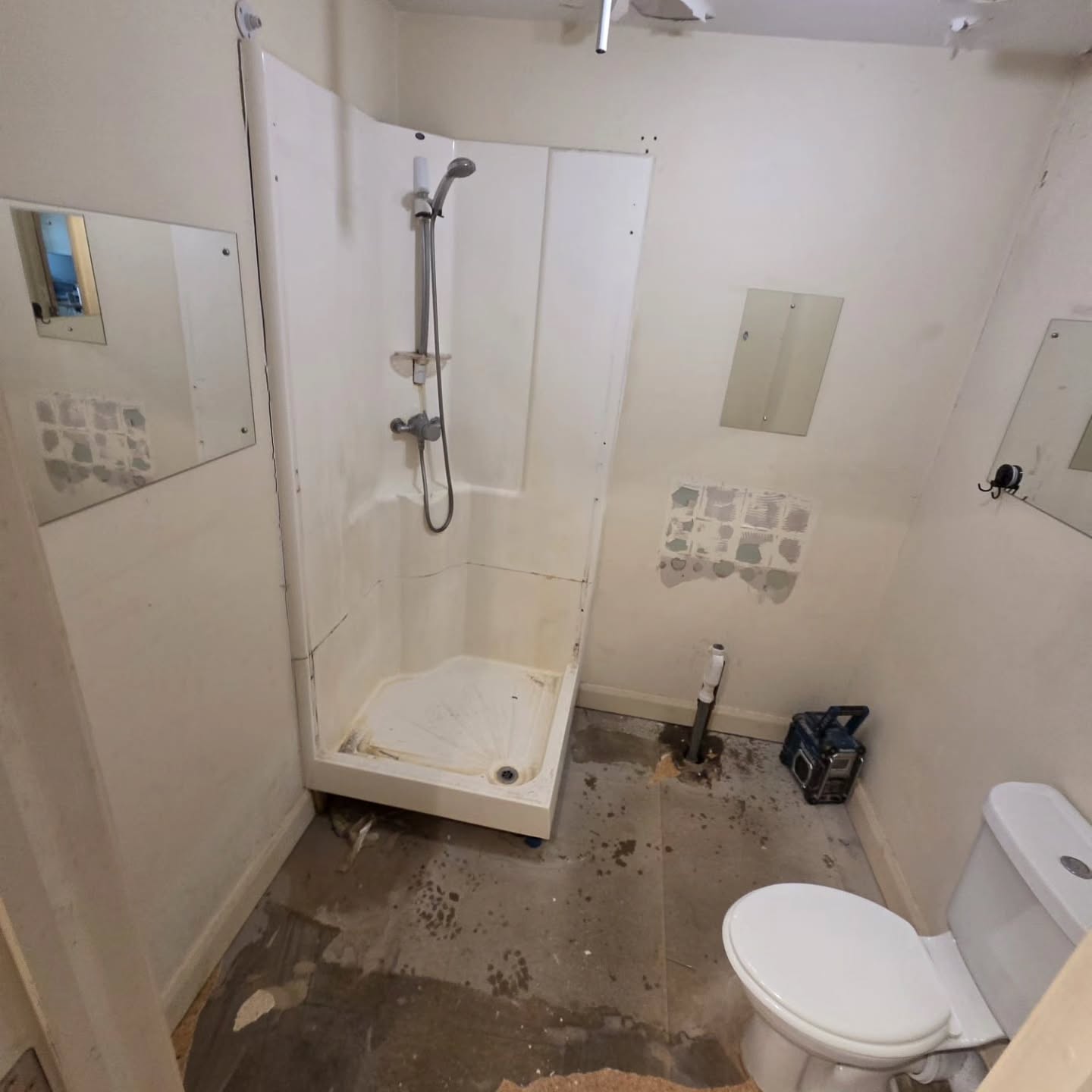 Before: tired bathroom mid-strip with old plastic shower enclosure, screw-fixed mirror panels, painted walls and bare concrete floor ready for renovation.